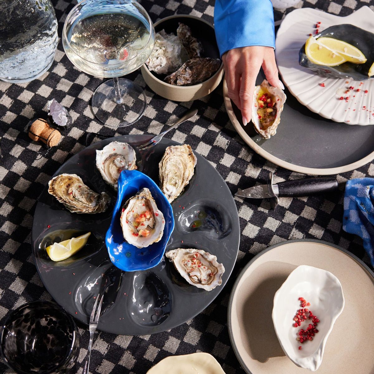 A hand serves an oyster beside a black oyster serving plate with oysters, lemon wedges, glassware, and tableware on a checkered table