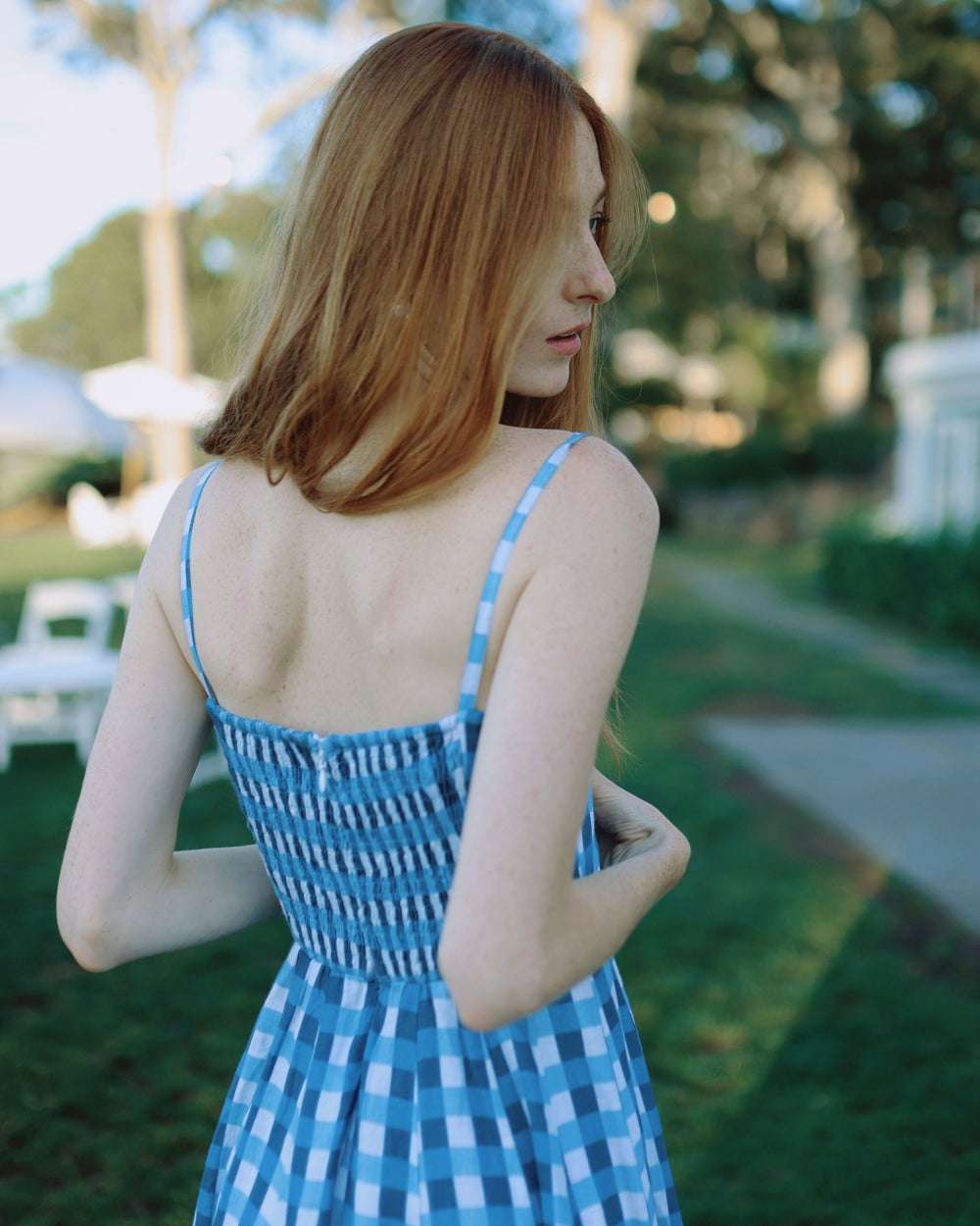 Back view of Allegra Dress in Cape Gingham with shirred bodice, thin straps, and blue gingham skirt outdoors