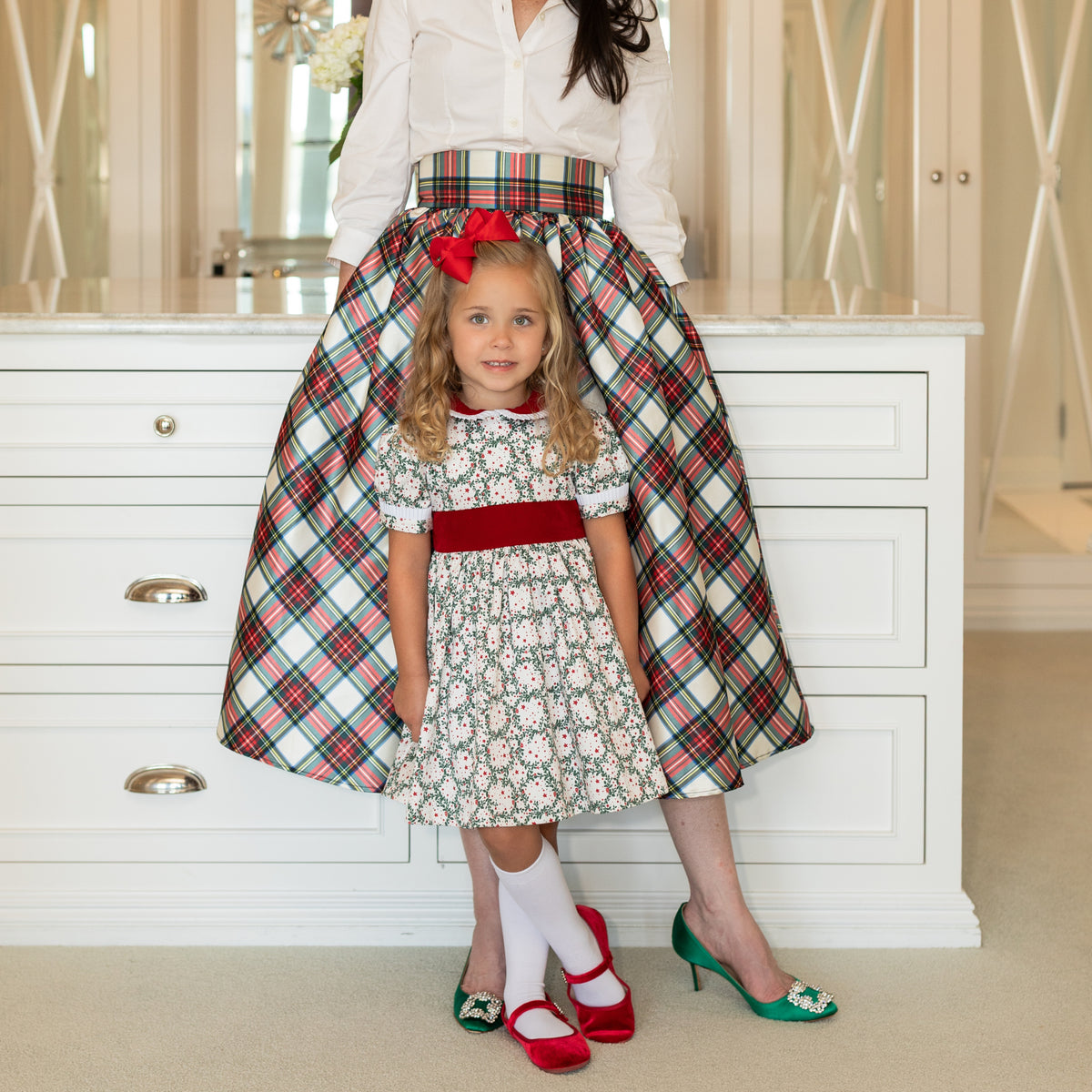 Front view of Short Sleeve Grace Dress in Regent Wreaths with red velvet collar, sash, and white pleating on a child in a kitchen