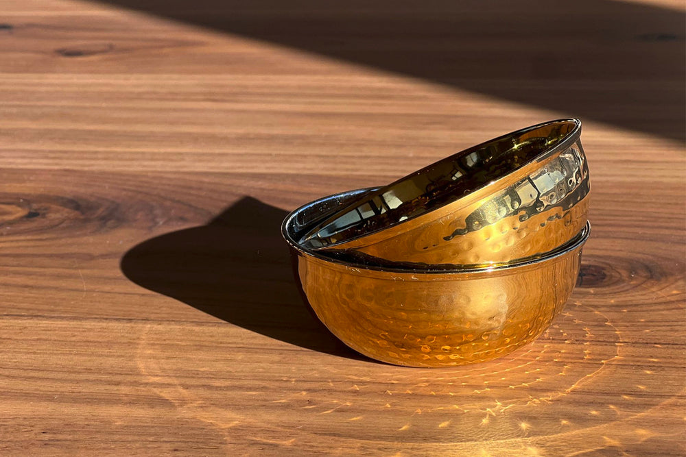 Side view of two stacked Dal serving bowls showcasing hammered stainless steel texture and warm gold reflections on a sunlit wooden surface