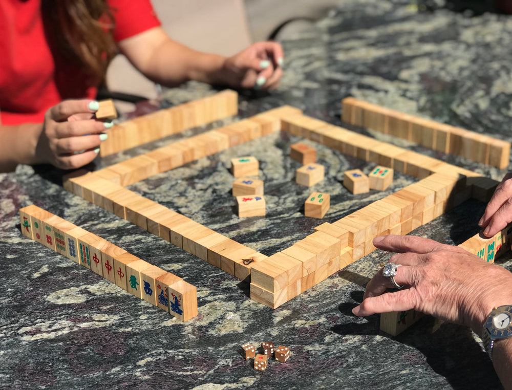 Two people playing Chinese mahjong with handmade wooden tiles and tile racks on a stone table, close-up of hands and dice