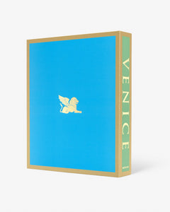 Venice: La Serenissima by Assouline: Three-quarter view of the back face and spine of the clamshell box. The back is solid sky blue with a small gold winged lion of Saint Mark emblem centered. The spine reads VENICE vertically on a green ground. Gold trim frames the box edges.