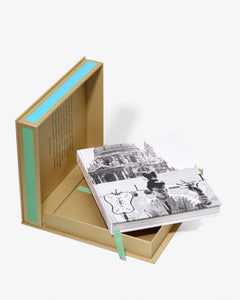 Venice: La Serenissima by Assouline: The open clamshell box with the book partially removed and resting at an angle in the tray. The cover shows a black-and-white vintage photograph of Venice — a masked Carnevale figure with gondolas and the columns of the Piazzetta in the background. A teal ribbon bookmark hangs below. Tan fabric lining with green accent stripes visible.