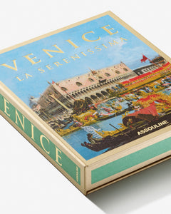 Venice: La Serenissima by Assouline: Angled overhead view of the closed clamshell box laid flat on a white surface. The box top shows the full Canaletto-style painting — VENICE in gold at top, LA SERENISSIMA below, ASSOULINE at lower center. The green-accented spine is visible at right with VENICE printed vertically.