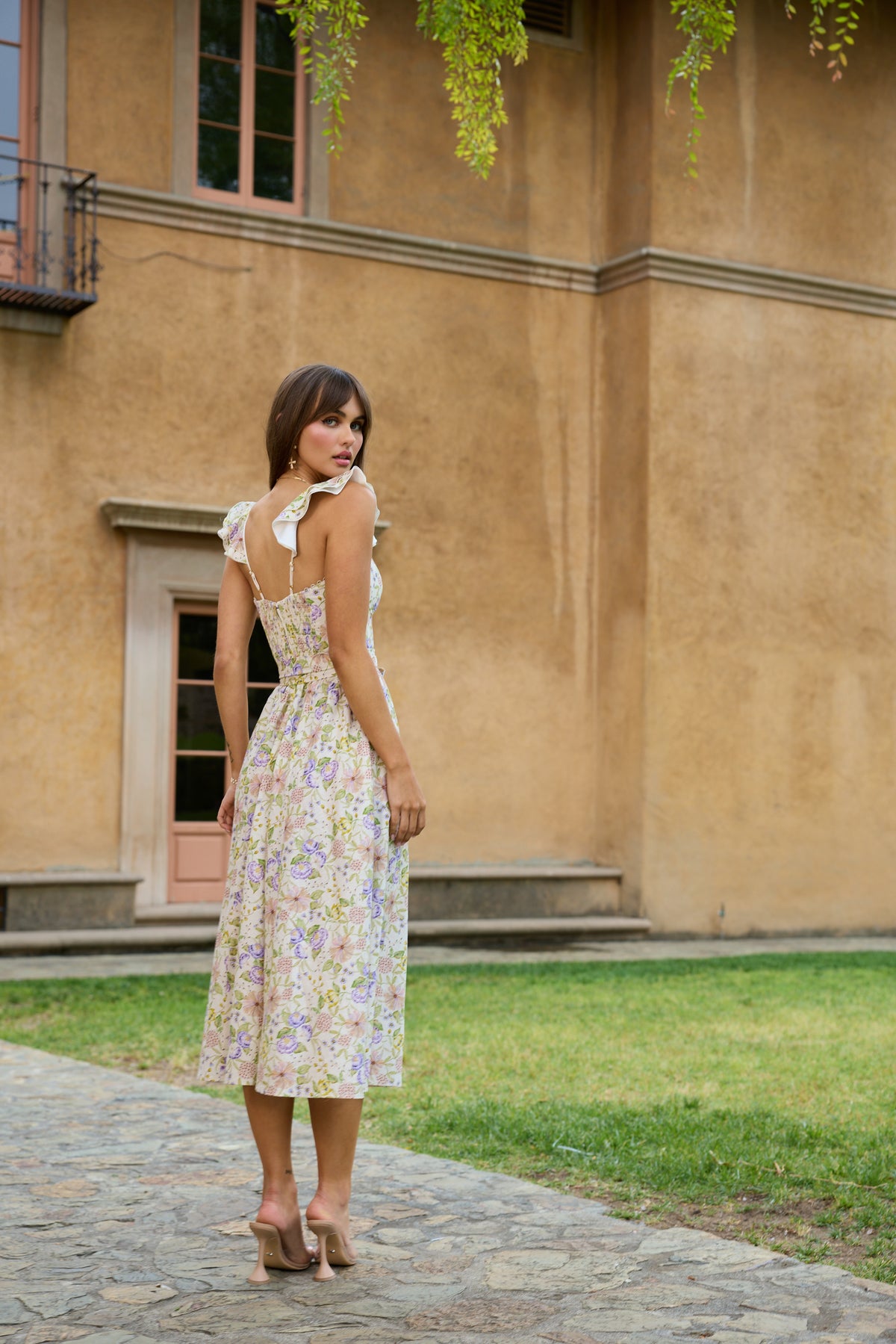 Back view of The Vera Dress in Cordoba Print showcasing its floral print and ruffle accents against a stucco building
