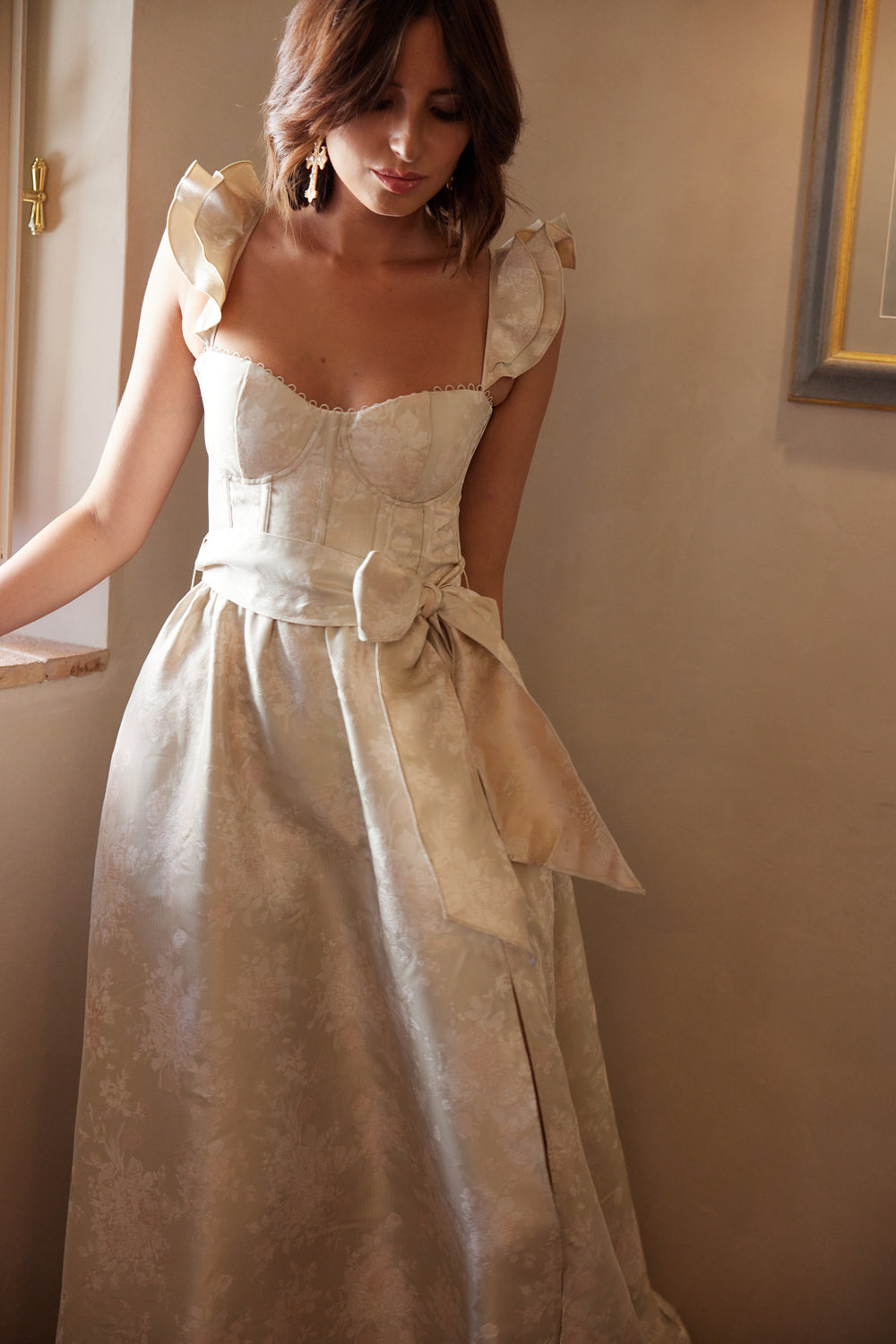 Three-quarter front view of Veronica Dress in taupe brocade with corset bodice, ruffled straps, bow waist, and side slit