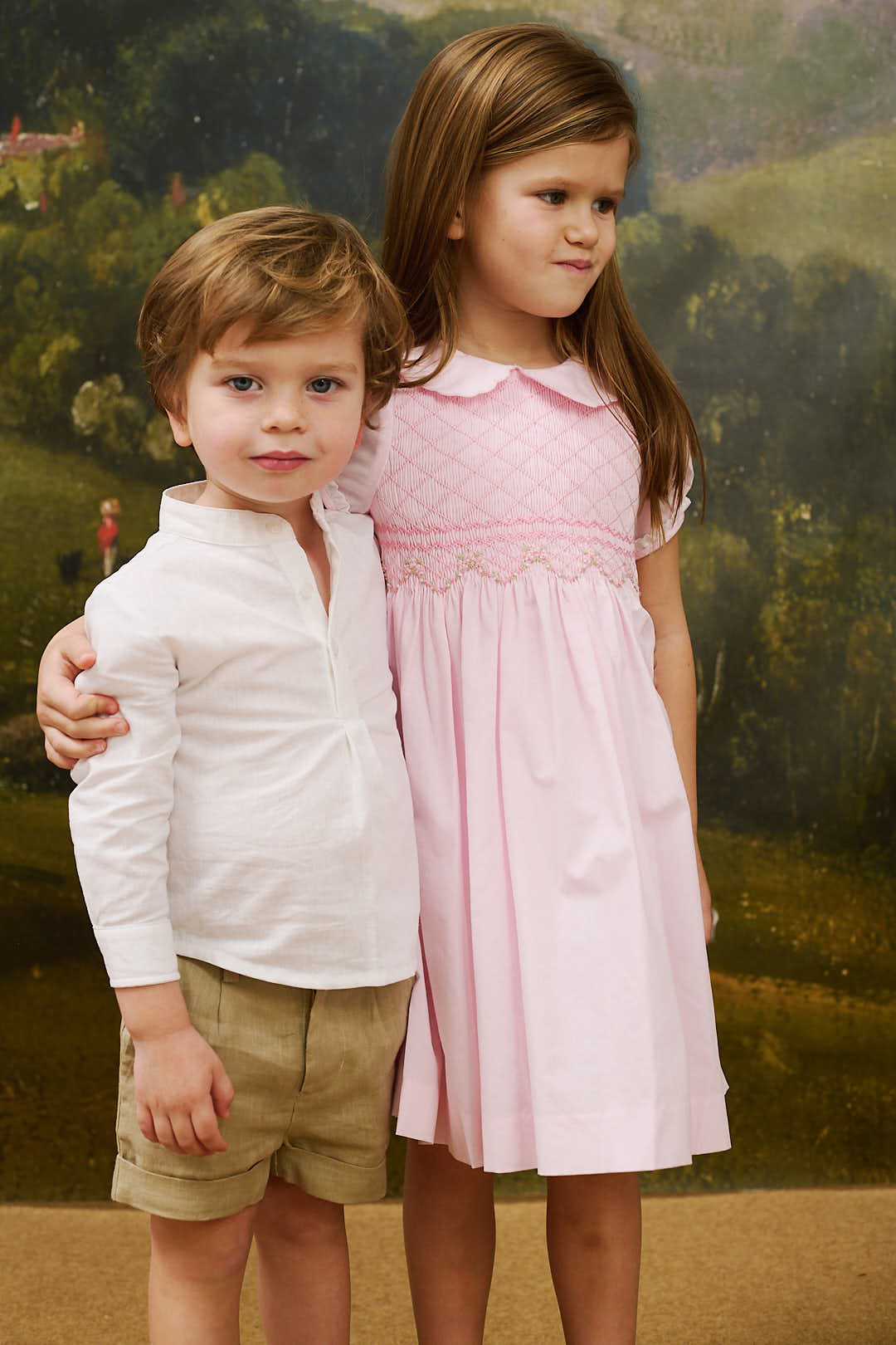 Boy wearing beige linen Jacobo shorts standing with a girl in a pink dress against a painted landscape backdrop