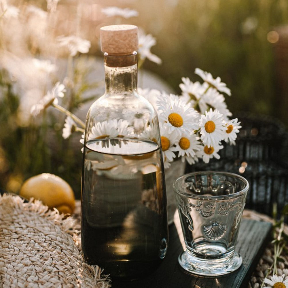 Front view of Versailles tumbler showing shell and floral motif and faceted skirt base on outdoor table with bottle and daisies