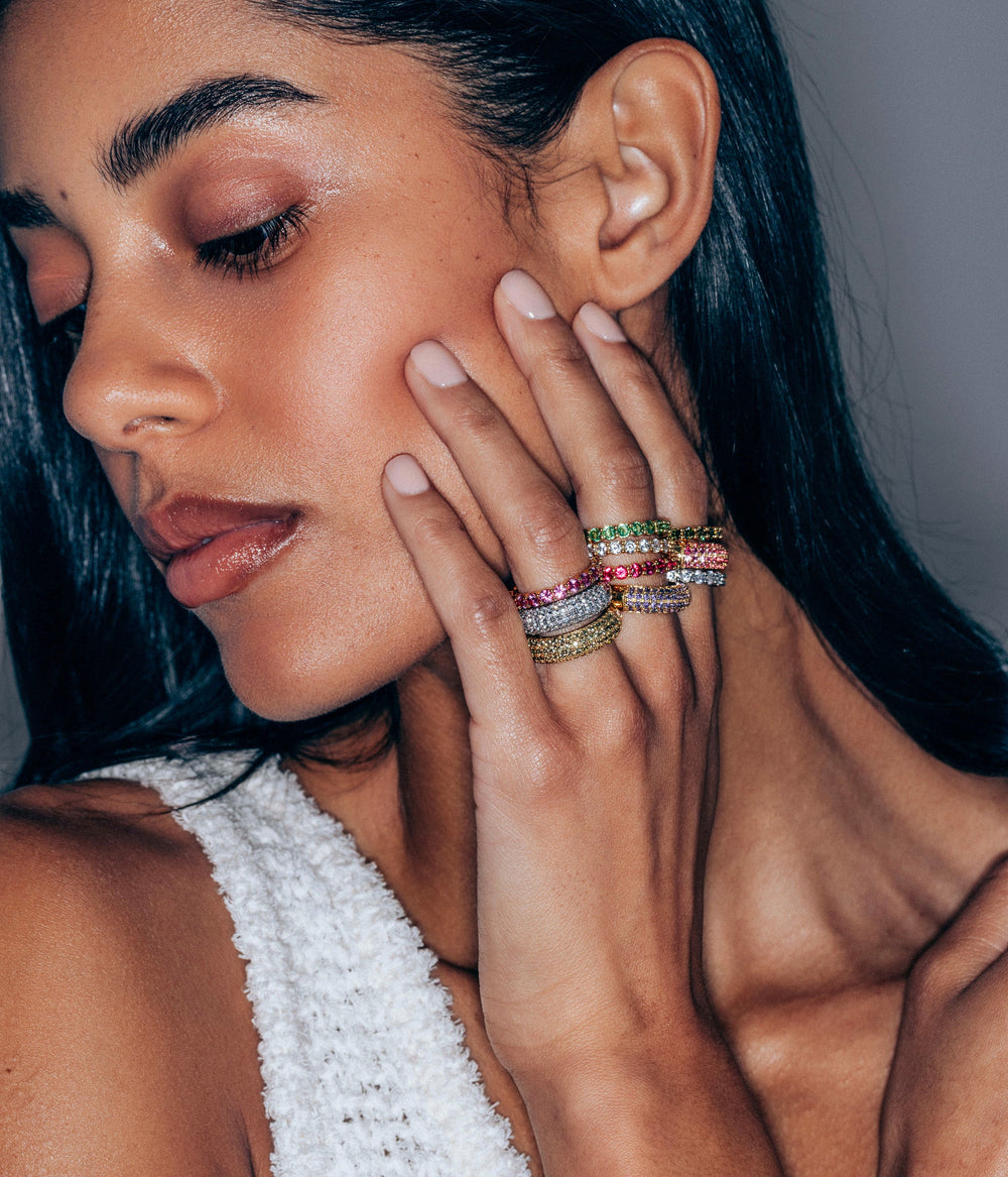 Close-up of hand wearing Veyra Ring in violet, pavé domed band stacked with colorful gemstone rings on gray background
