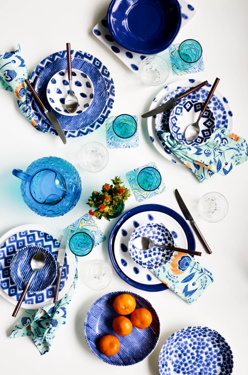 Overhead table setting with blue-and-white Santorini patterned plates and bowls, glasses, cutlery, napkins, and oranges