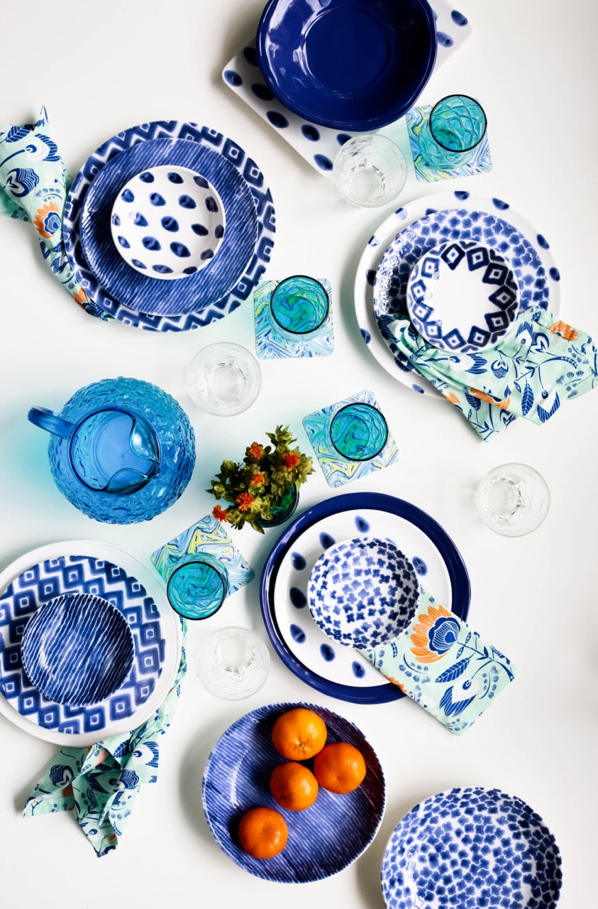 Overhead view of blue-and-white Santorini plates and condiment bowls with glasses, patterned napkins, and oranges on a white table