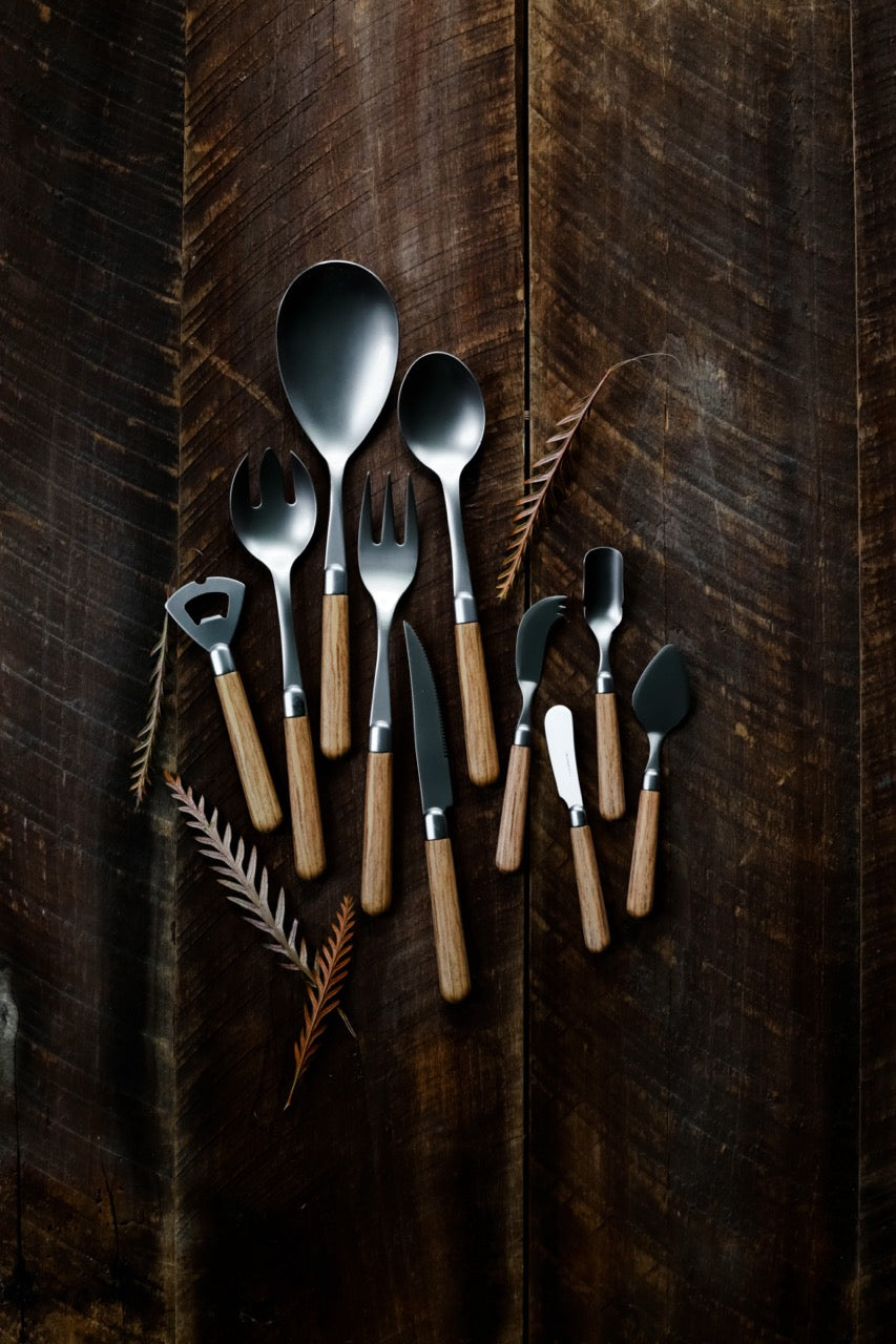 Top-down view of Albero spreader and serving utensils showcasing wood-toned handles and matte dark stainless blades on dark wood background