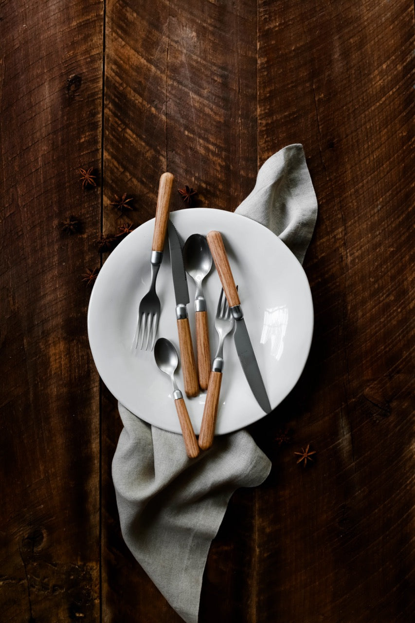 Top-down view of Albero five-piece place setting with wood-handled stainless utensils on a white plate over dark wood