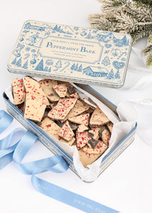 Open Tuck Shop for Over The Moon Peppermint Bark tin showcasing peppermint-topped bark pieces with blue ribbon and evergreen branches