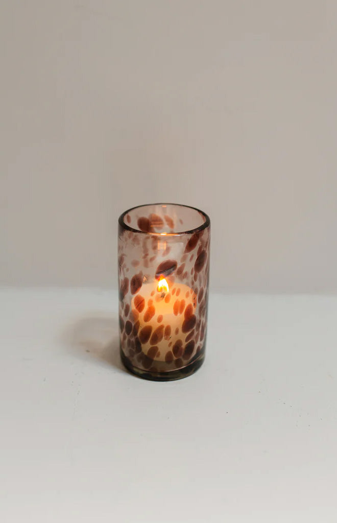 Front view of a single amethyst-marble glass votive with lit candle showing elongated silhouette and purple marbled pattern on white background