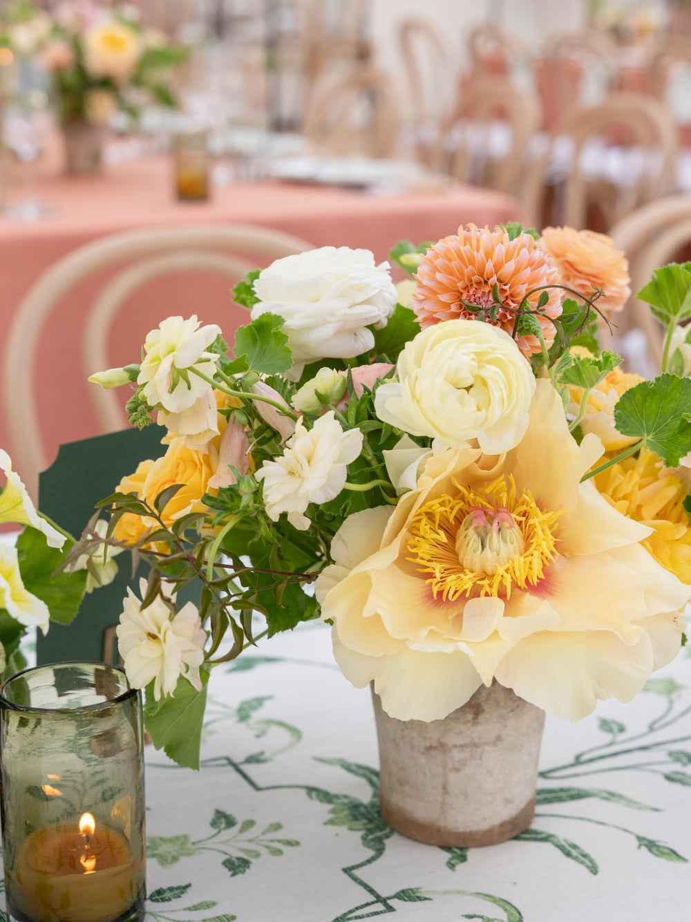 Lit light-olive glass votive candle beside a peach-and-yellow floral centerpiece on a decorated dining table with bentwood chairs in the background