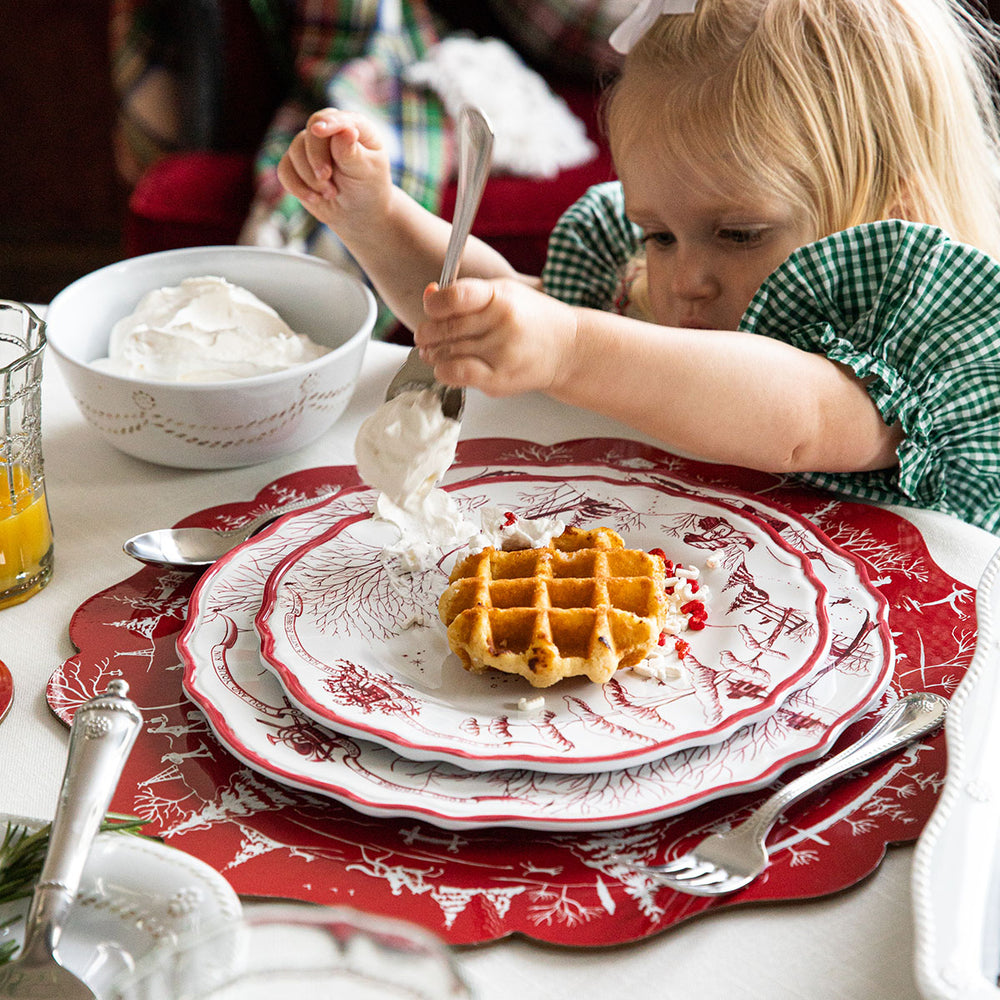 Child spooning whipped cream onto a waffle on stacked red-and-white Winter Frolic melamine plates at a holiday table