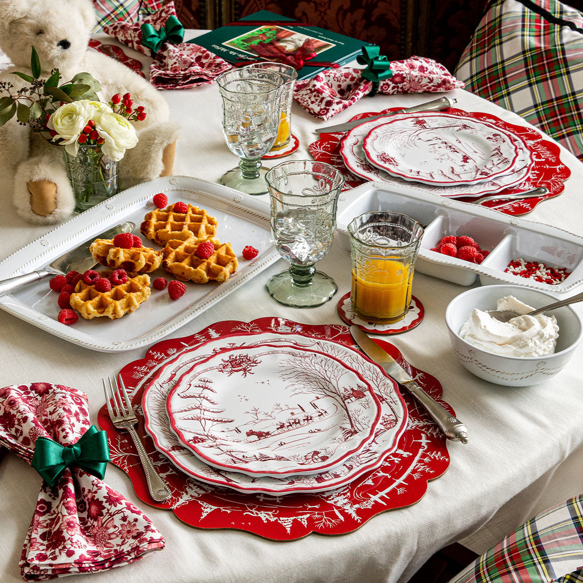 Stacked red-and-white Winter Frolic salad plates on a holiday table with waffles, glassware, and a napkin tied with a green ribbon
