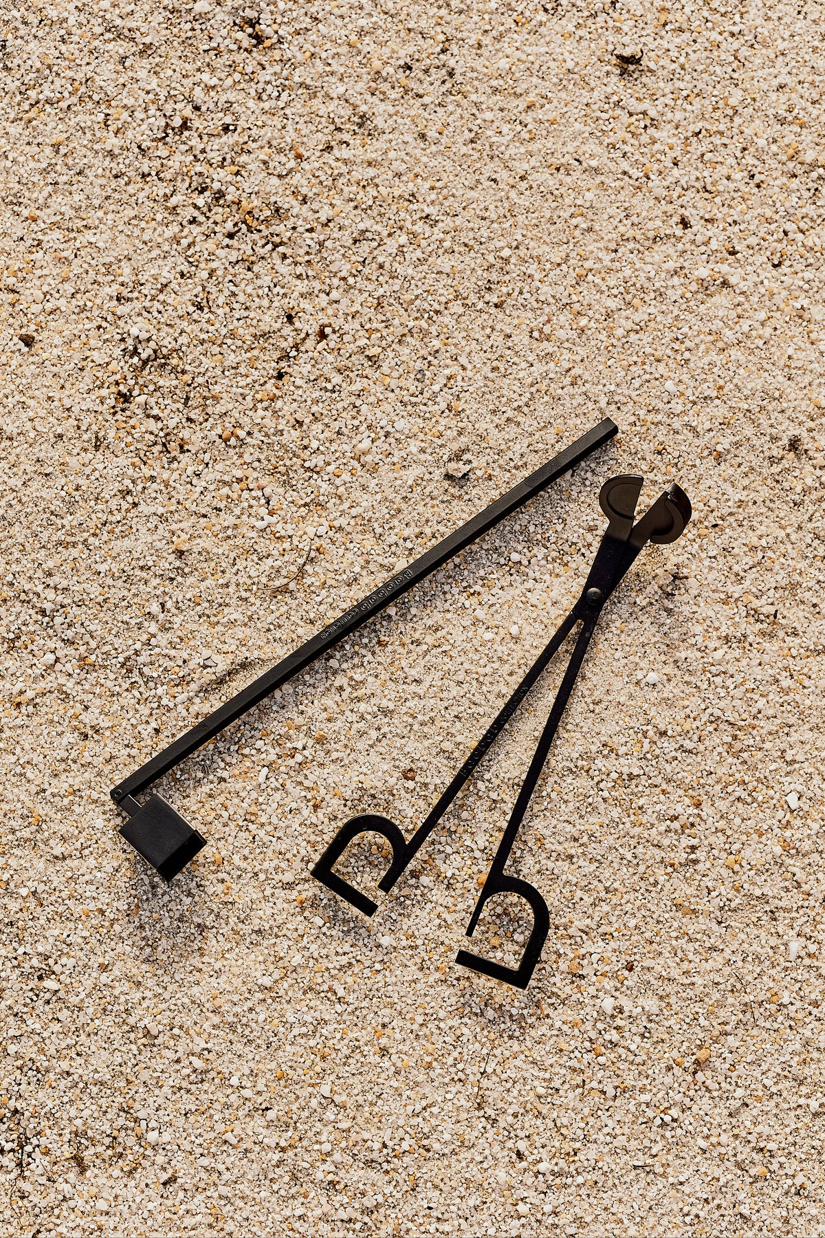 Angled top view of matte black metal wick trimmer showing scissor-style snips and long handle on sandy background