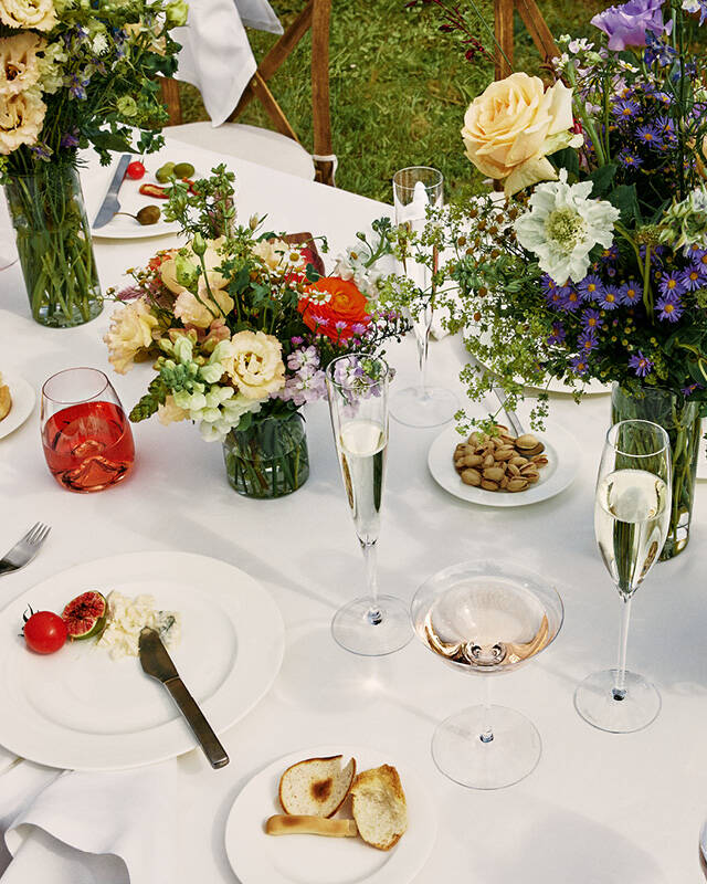 Crystal champagne flutes and a coupe arranged on a white-linen table set for a garden meal with flowers and plates