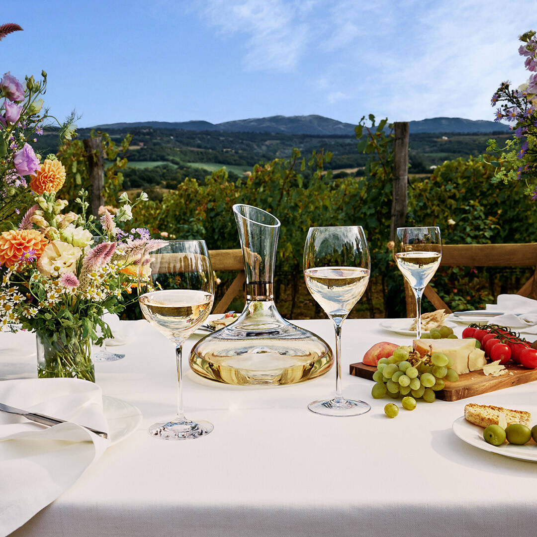 Angled view of Elegance Bordeaux wine glasses set with tapered bowls and slender stems on a table with decanter and grapes