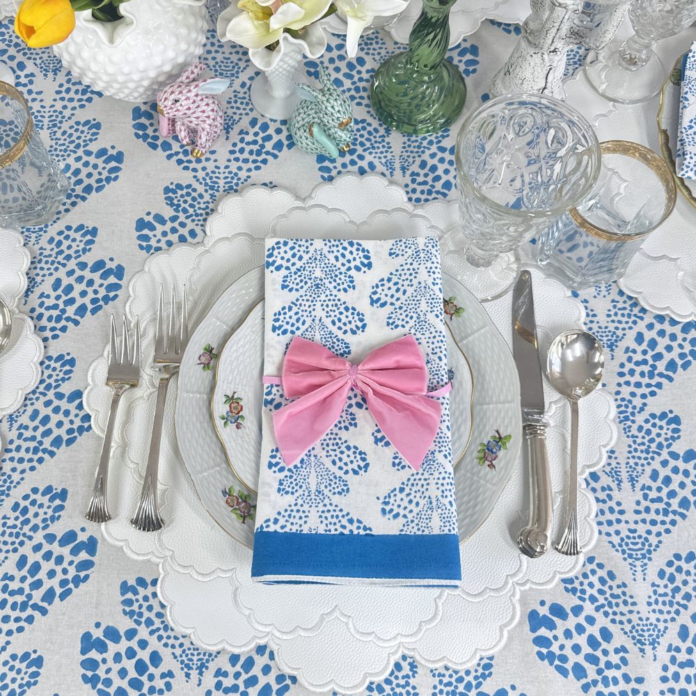 Dining table setting with floral tablecloth, plates, cutlery, and a pink bow on a napkin. white vegan leather luxury placemat. 