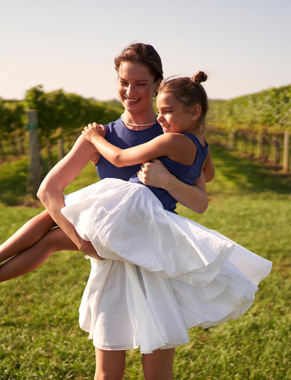 Girl wearing white Mini Willa tutu skirt and navy top being carried by a woman in a sunlit vineyard