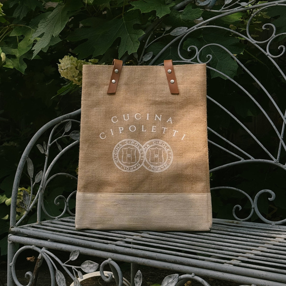 A beige wine tote with leather straps and the 'Cucina Cipoletti' logo printed on the front, placed on a metal bench with foliage in the background.