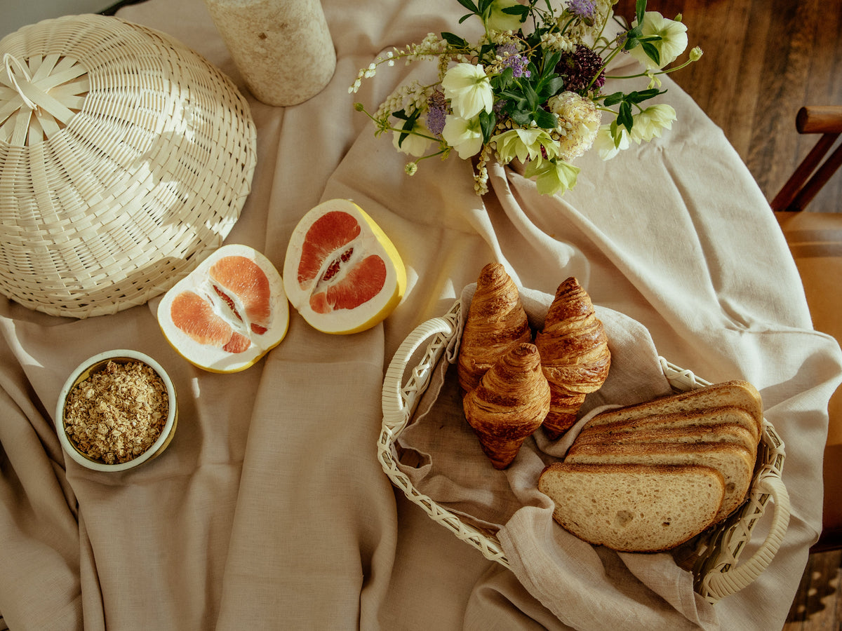 Folk Fleur cloche beside a basket of croissants and sliced bread on a beige linen-covered table with halved grapefruit, a bowl of granola, and wildflowers
