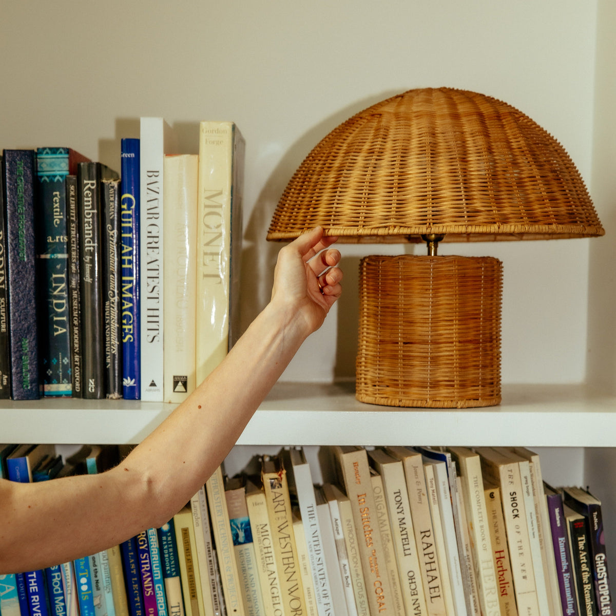 Hand adjusting woven rattan Lanai Table Lamp on a white bookshelf with stacked books