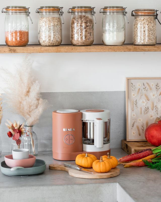 Three-quarter view of Babycook Neo food maker in terracotta showing glass bowl and stainless-steel steam basket on a kitchen counter