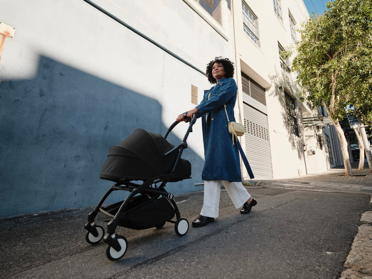 Woman pushing a black YOYO3 stroller with newborn pack along a sunny urban sidewalk
