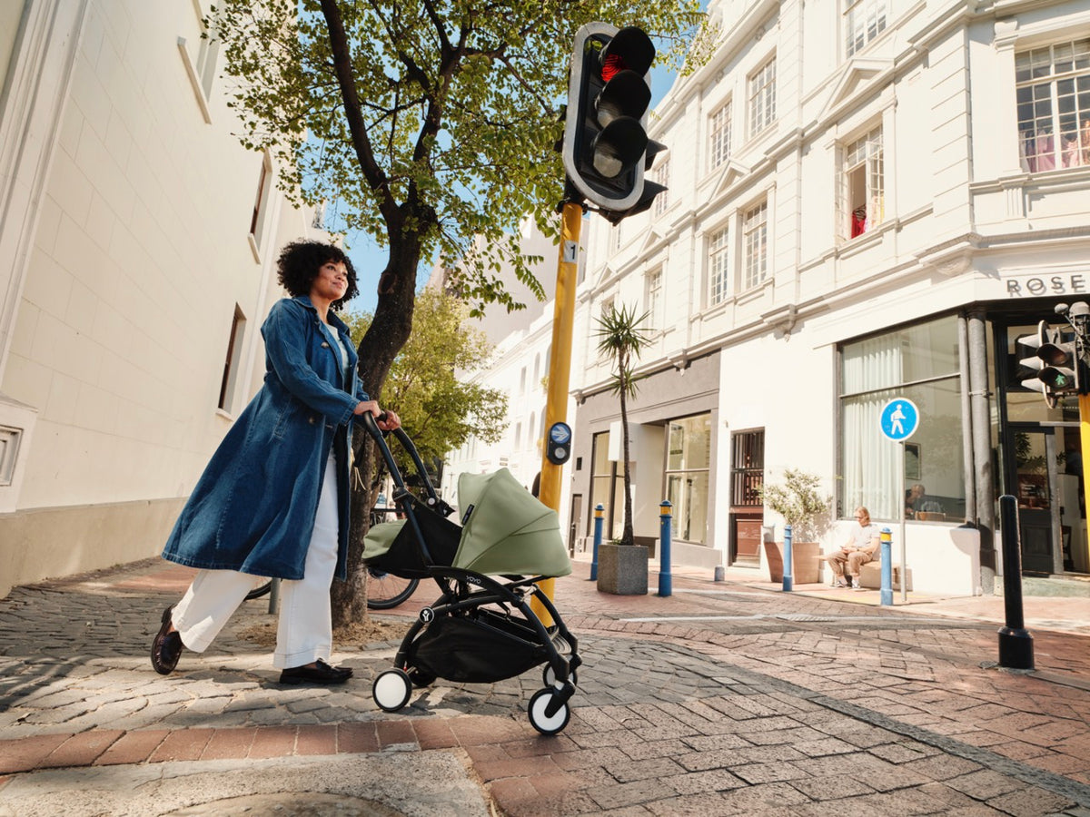Woman pushing a YOYO³ stroller fitted with Olive 0-plus Newborn Pack along a sunny city sidewalk