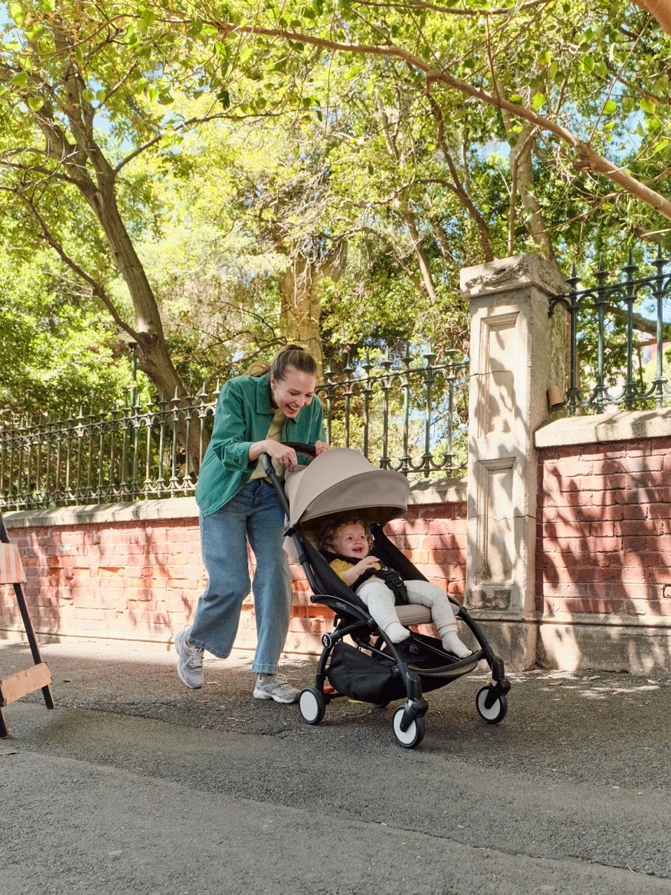 Woman pushing a toddler in a YOYO³ stroller with black frame and taupe canopy along a tree-lined sidewalk