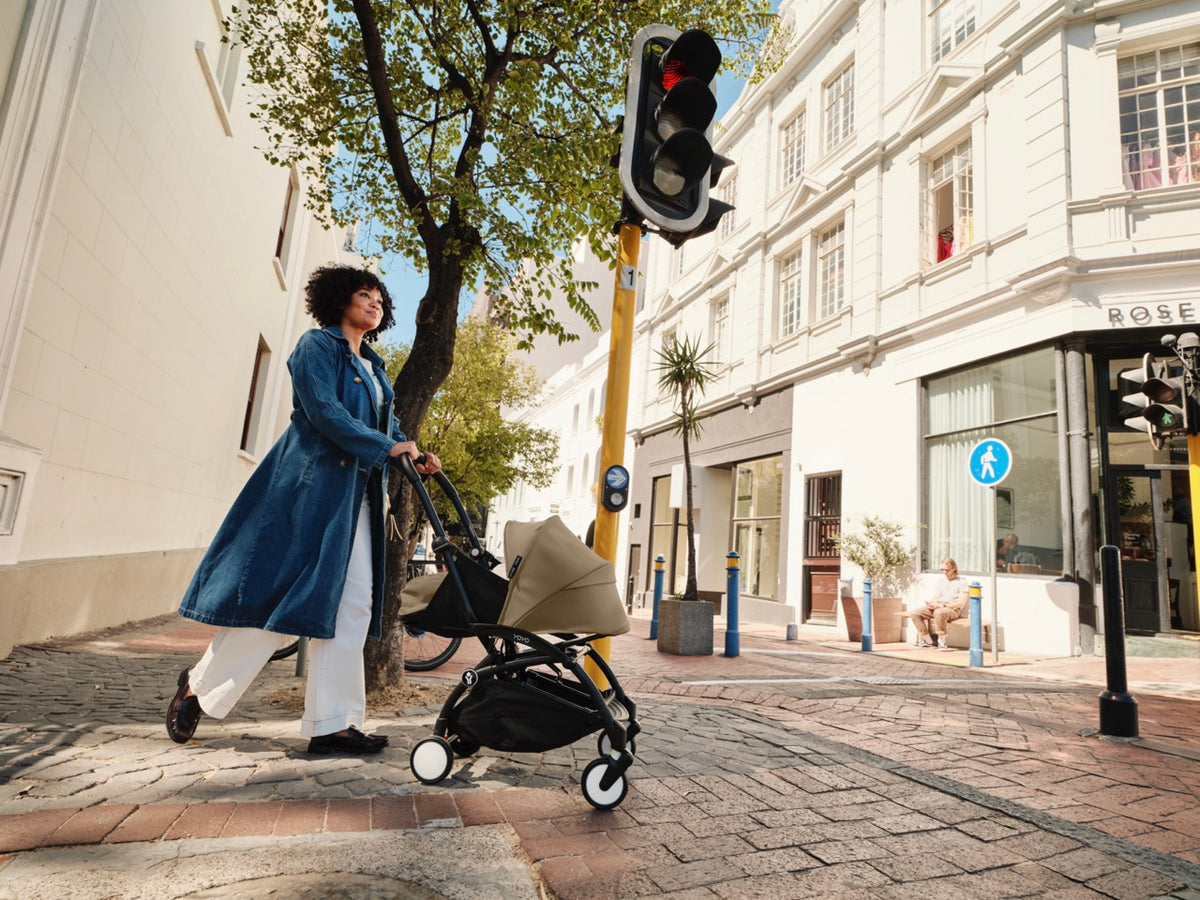 Woman pushing a Toffee YOYO³ stroller on a sunlit city sidewalk past a traffic light