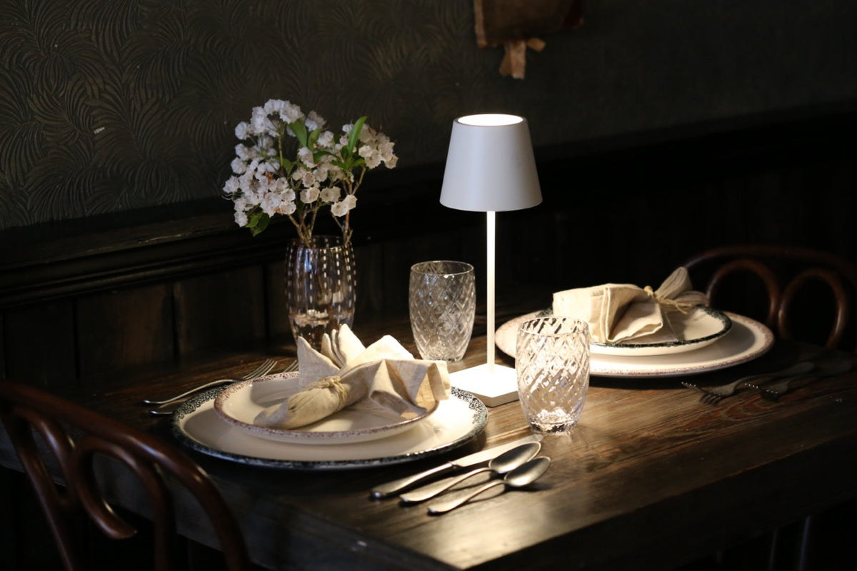 Poldina Pro Mini table lamp illuminating a rustic two-place tablescape with plates, cloth napkins, crystal glasses, and a small vase of flowers