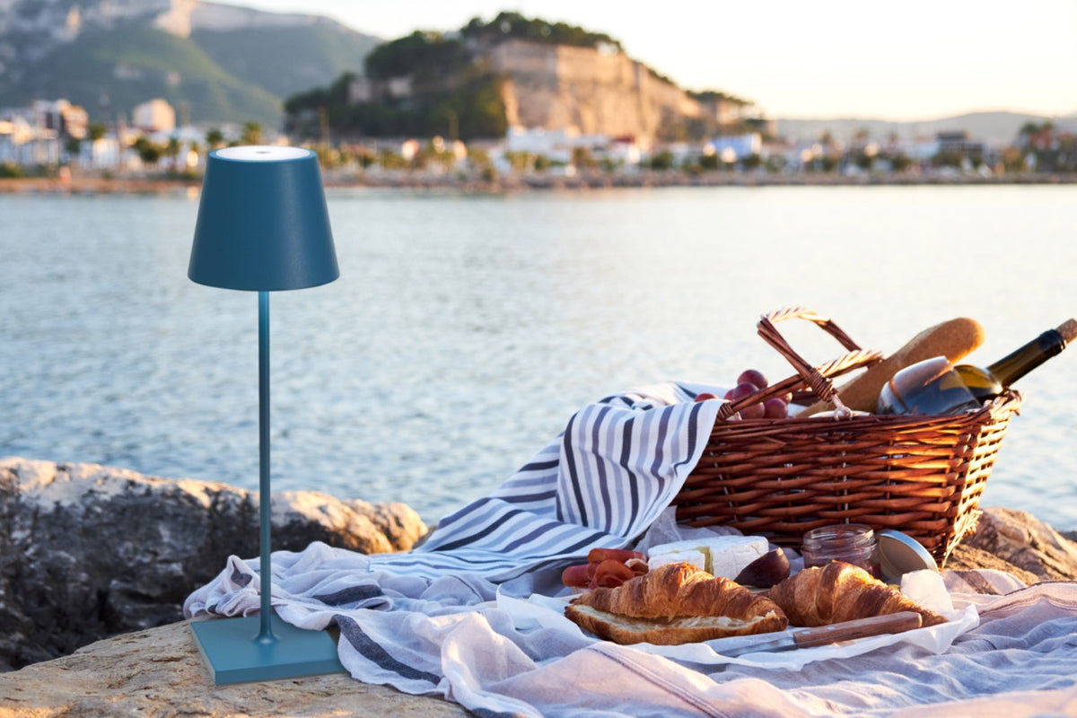 Upright Poldina Pro table lamp showing slim stem and blue shade on a picnic blanket by the seaside