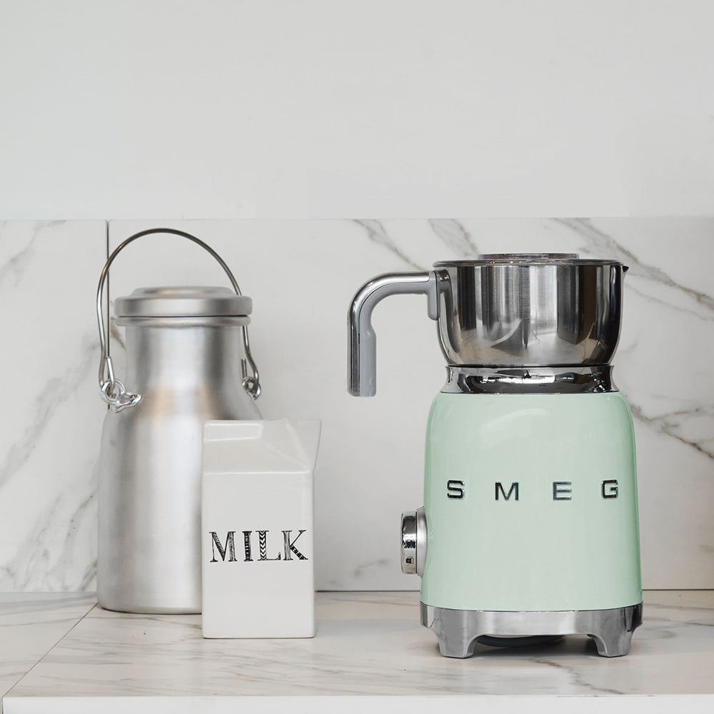 Front view of SMEG milk frother with stainless steel jug and pastel green base on marble countertop beside milk can