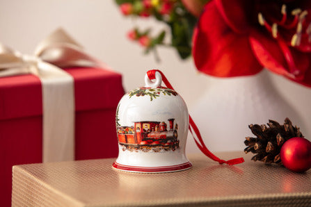 Close-up front view of porcelain bell ornament showcasing a hand-painted train scene, gold trim, and red ribbon on a wrapped gift