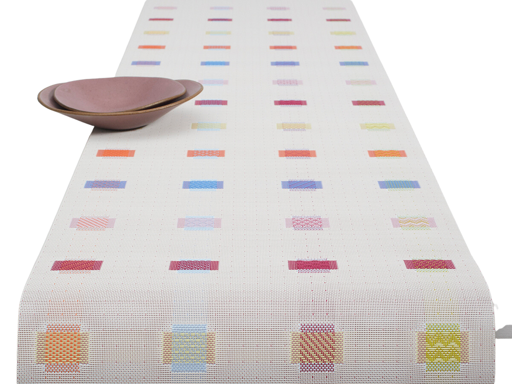 Angled top view of Sampler table runner showing multicolored stitched rectangles on white woven ground with two mauve bowls