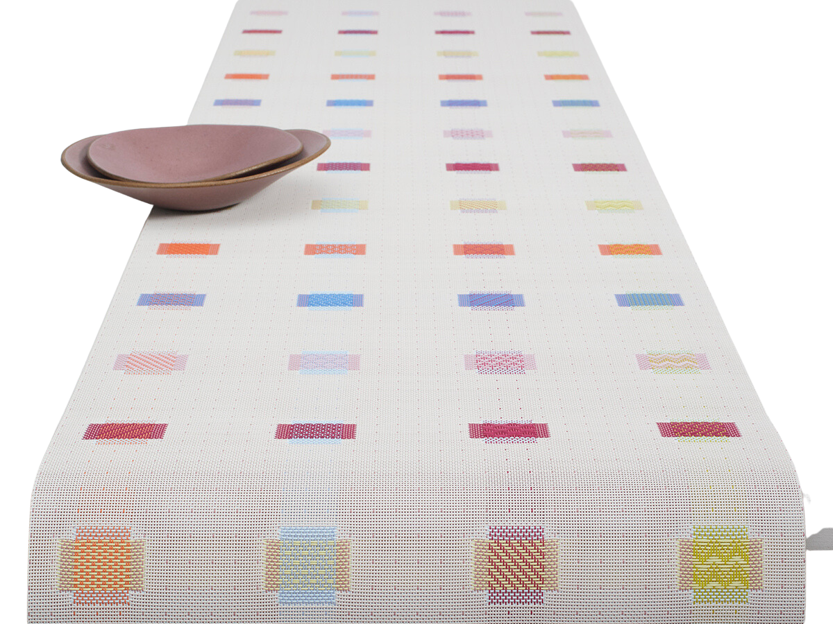 Angled top view of Sampler table runner showing multicolored stitched rectangles on white woven ground with two mauve bowls
