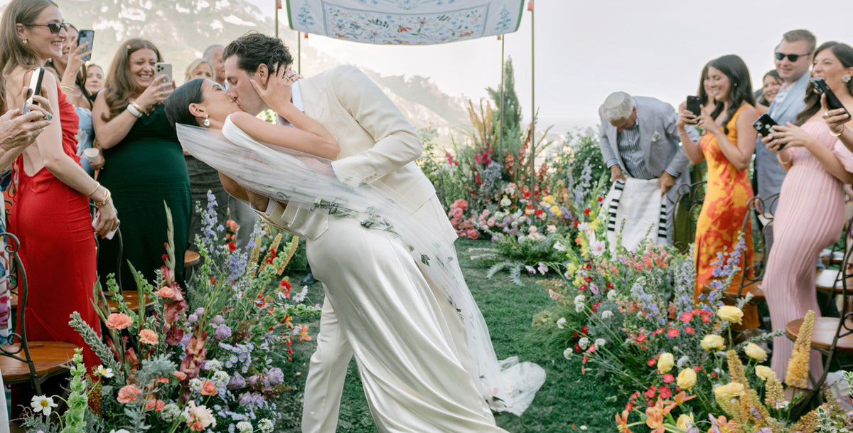 Newlywed couple kiss during an outdoor wedding ceremony surrounded by colorful flowers and photographing guests in Ravello