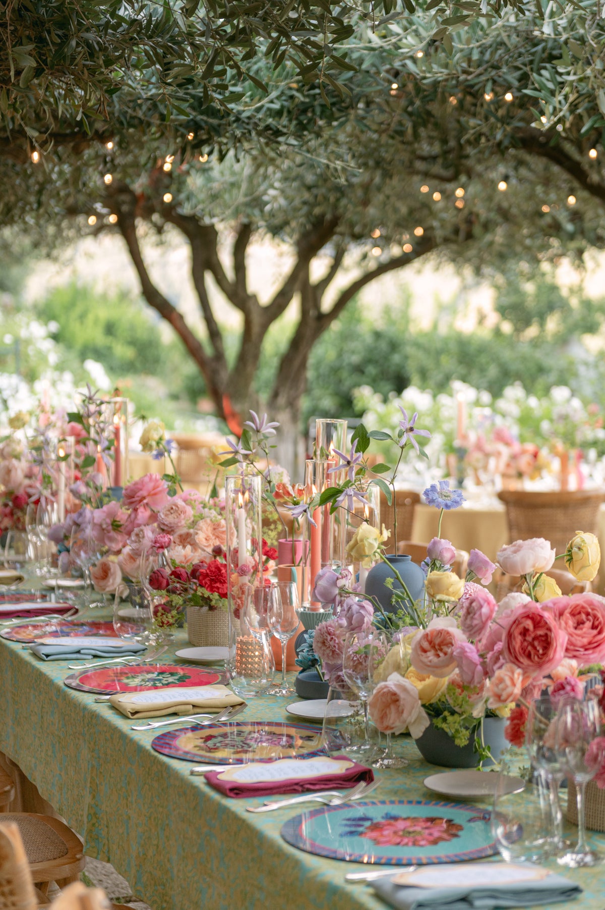 Outdoor wedding dinner table set beneath an olive tree with colorful flowers, candles, and patterned plates