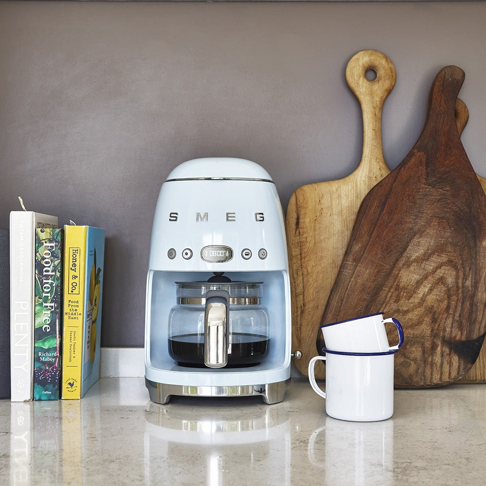 Front of SMEG drip coffee machine showing glass carafe and chrome accents on countertop with cutting boards and enamel mugs