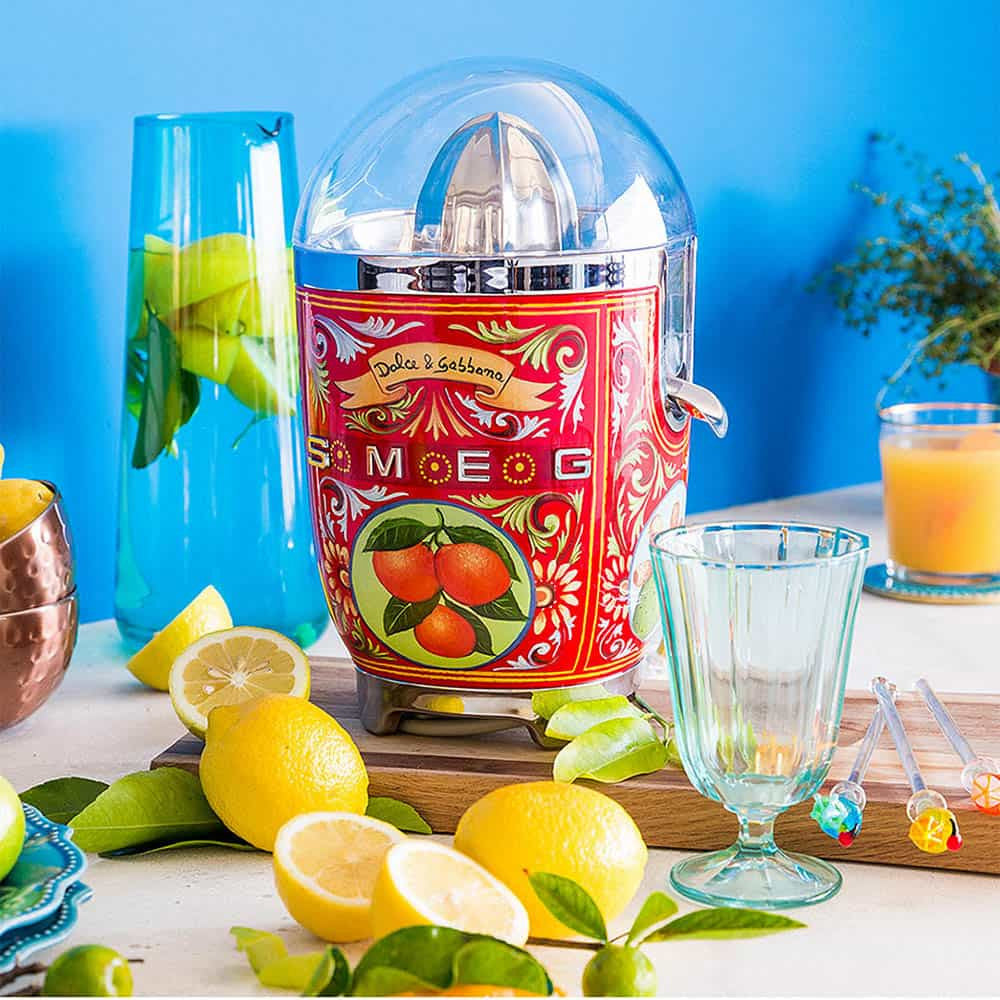 Front view of SMEG x Dolce and Gabbana citrus juicer with red fruit design and clear dome on blue tabletop with lemons