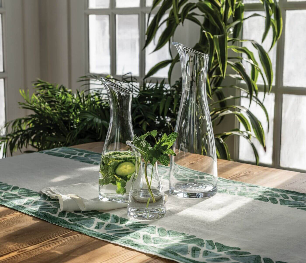 Three Simon Pearce Barre carafes on a sunlit wooden table with a green leaf table runner and houseplants in the background