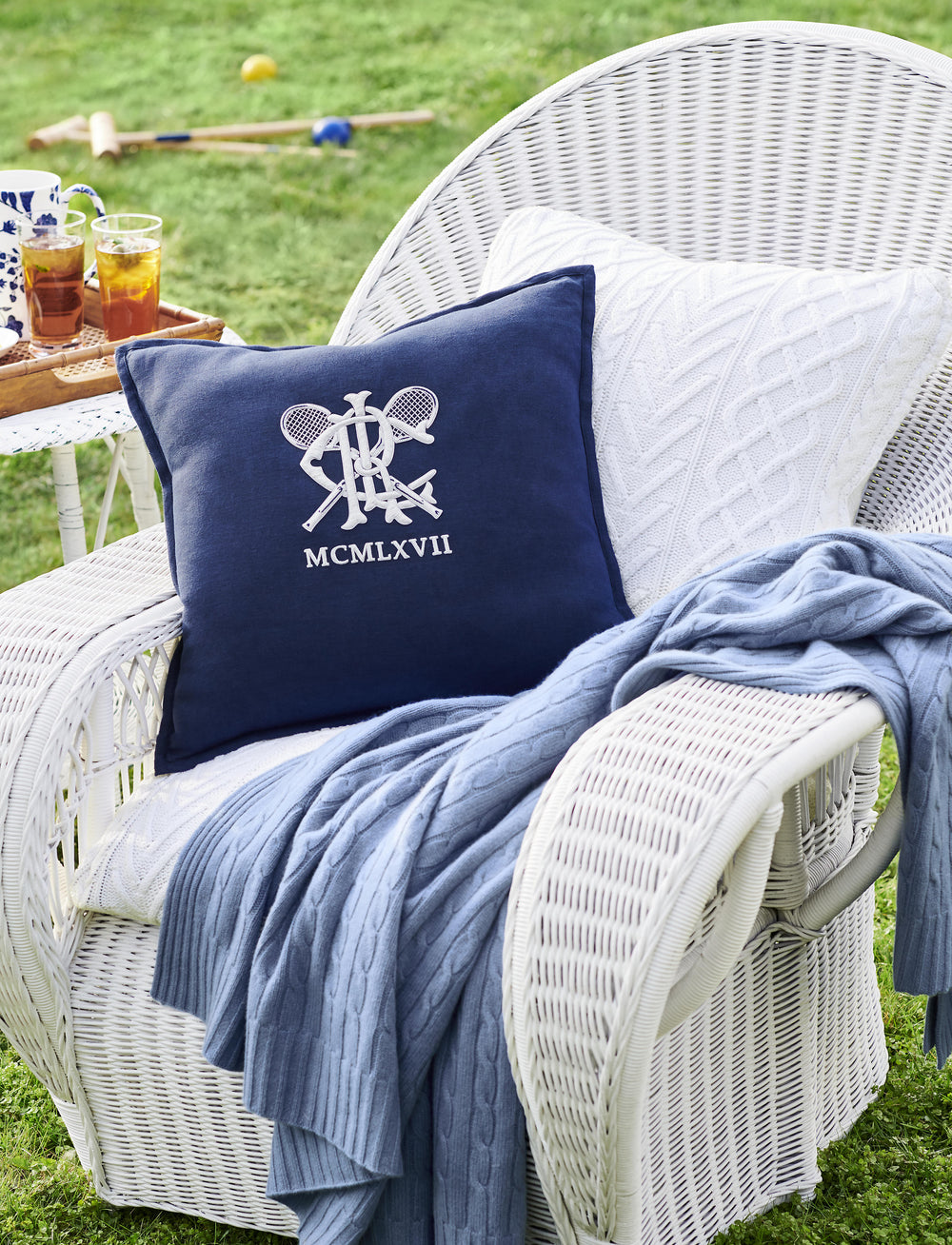 Heathered blue cable cashmere throw draped over white wicker chair with navy monogram pillow on a sunny lawn