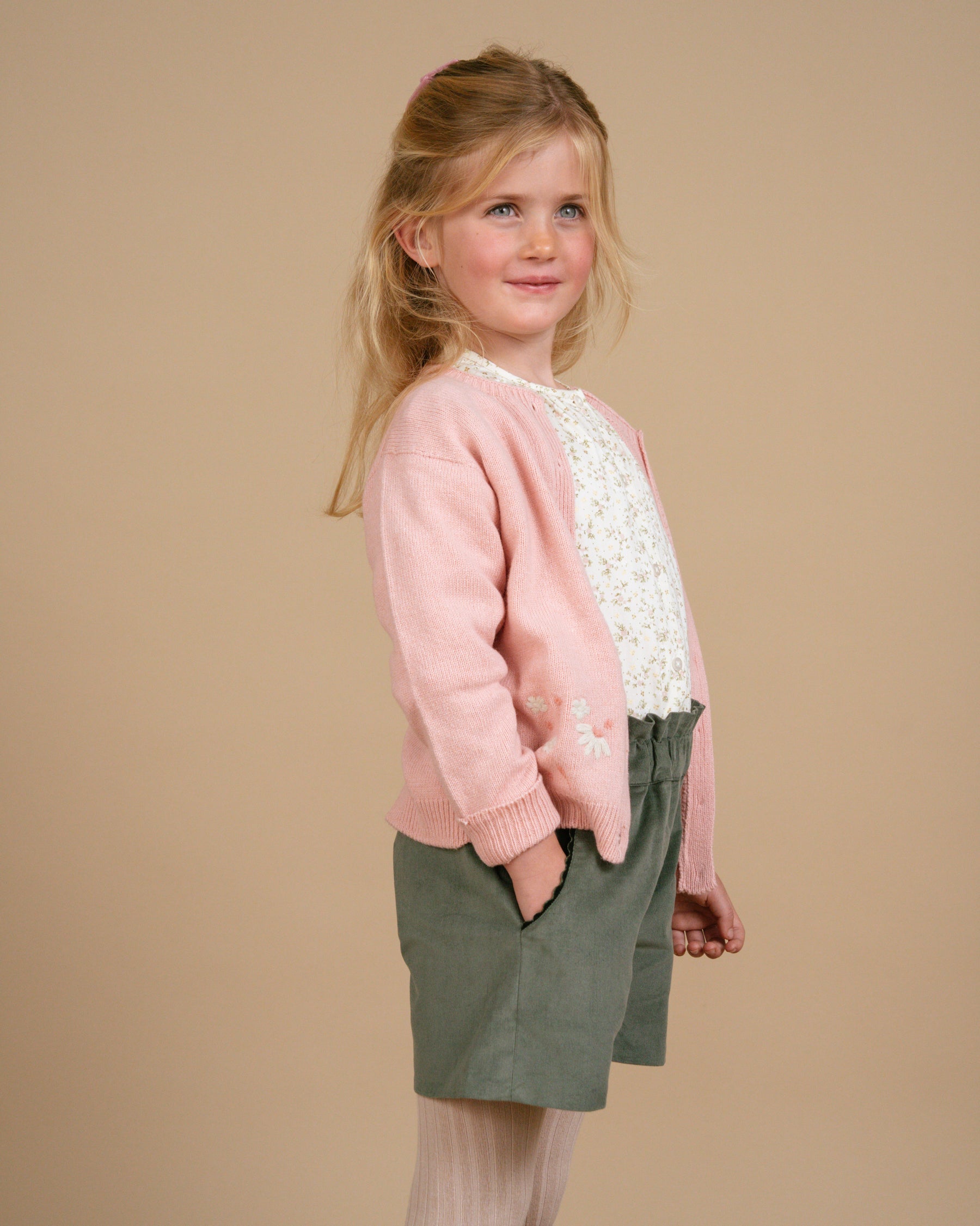 Young girl wearing a pink sweater with lace details and green shorts on a beige background