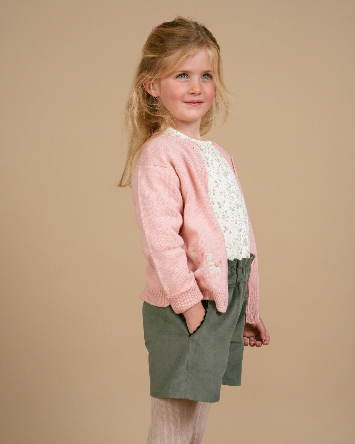 Young girl wearing a pink sweater with lace details and green shorts on a beige background