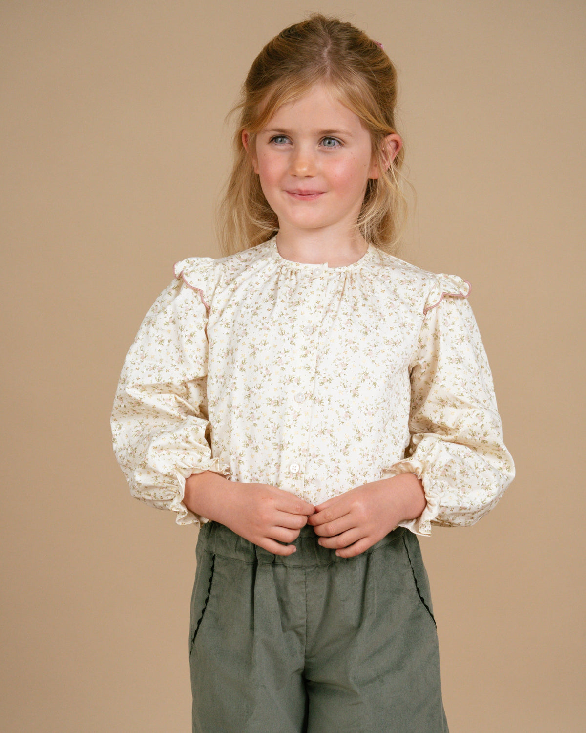Young girl wearing a white lace blouse and green pants against a beige background